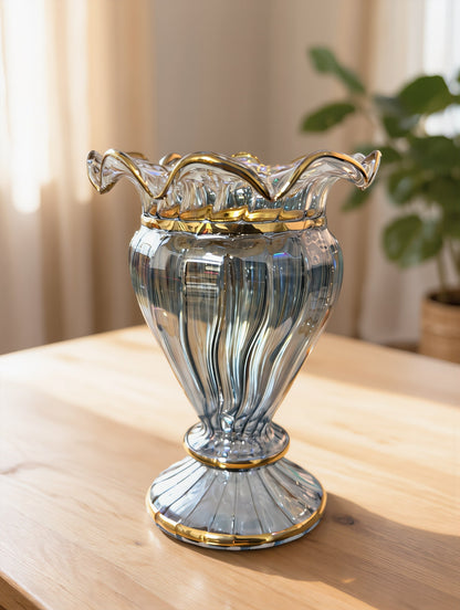 A ribbed clear glass vase with a flared rim and gold trim, set on a light wood table in a softly lit room with a blurred plant in the background.