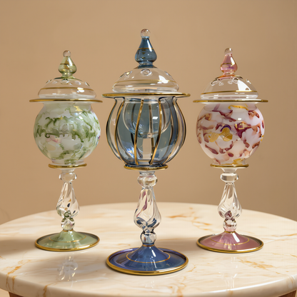 Three lidded Egyptian glass mini jars in green, blue and pink with gold detailing on a white table