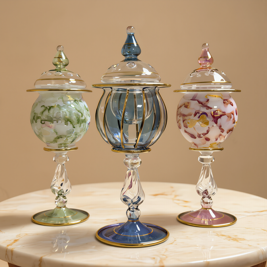 Three lidded Egyptian glass mini jars in green, blue and pink with gold detailing on a white table